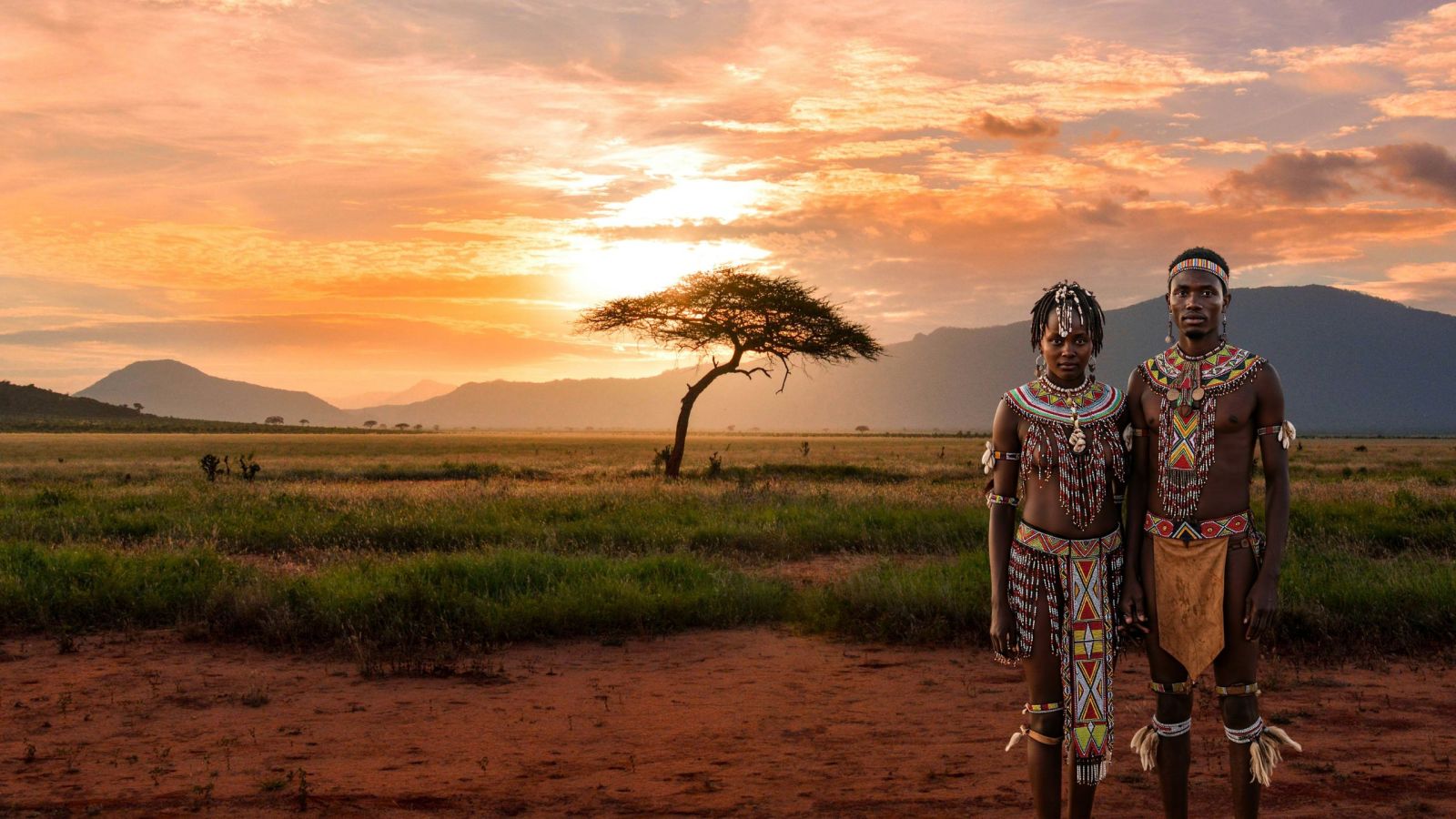 Ein Khoisan-Paar steht in einem offenen Feld während eines afrikanischen Sonnenuntergangs. Die Frau praktiziert Schamlippenverlängerung, gekleidet in traditioneller Kleidung, mit einem Baum, der im Hintergrund silhouettiert ist.